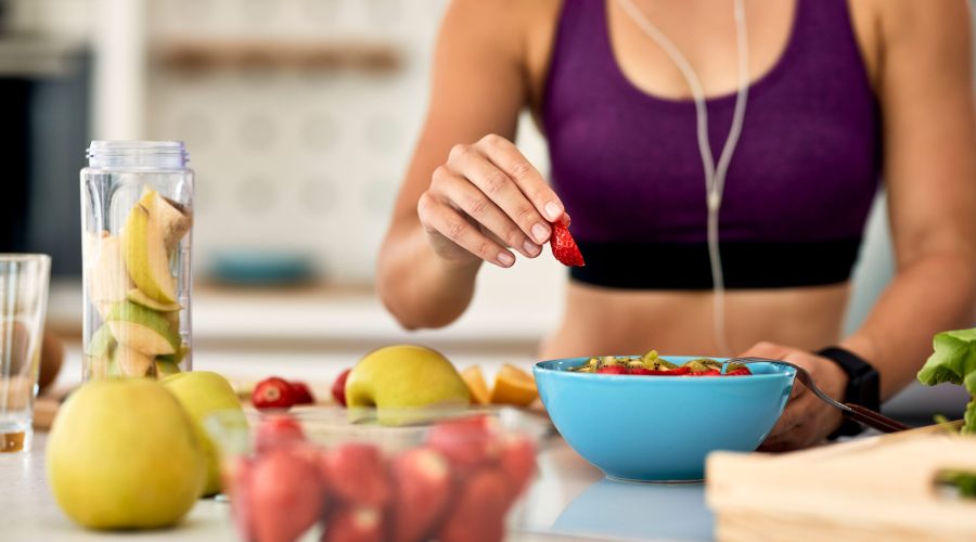 Close-up of athletic woman adding strawberries while making fruit salad in the kitchen.